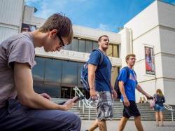 Two students walking past the Student Center and a third sitting looking at his phone
